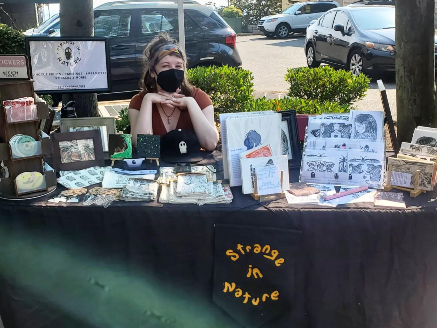 A black table with the logo Strange in Nature in front of it. Displayed on the table are several hand-printed items made by Krista O'Halpin, the owner of Strange in Nature. She is seated happily behind the table, but is wearing a black medical